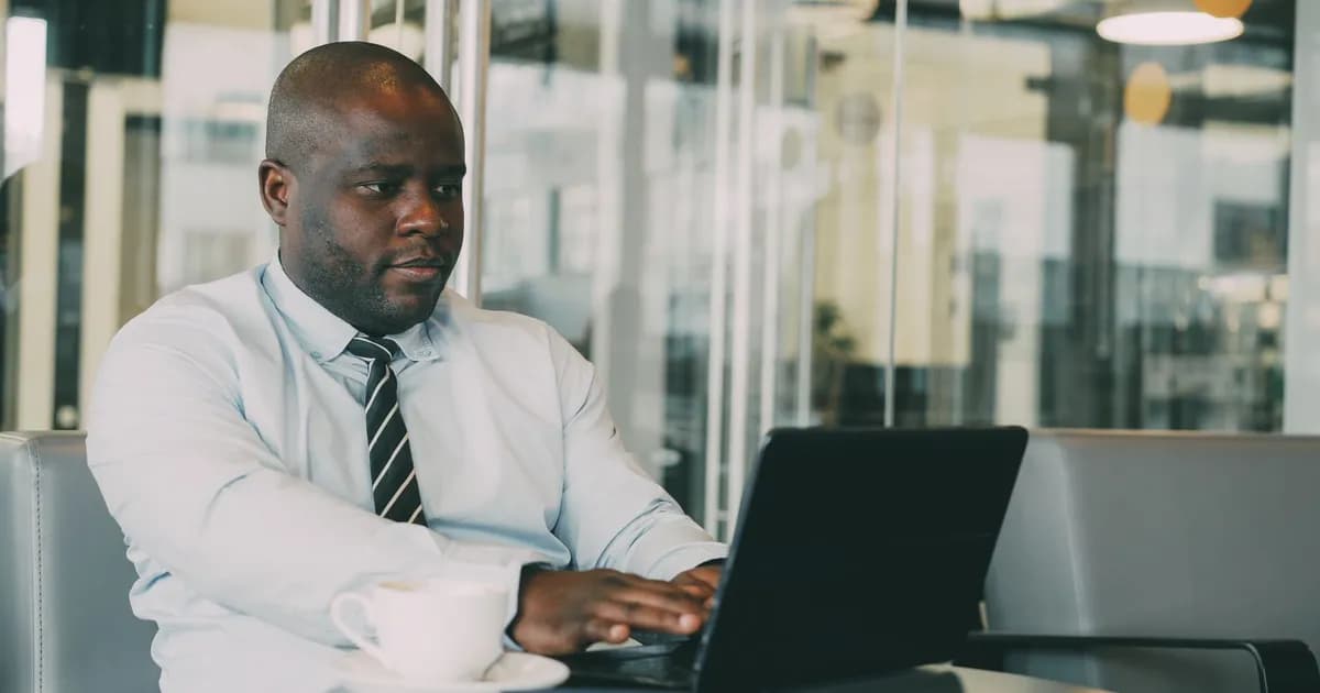 Man in a light blue shirt and striped tie working on a laptop beside a coffee cup