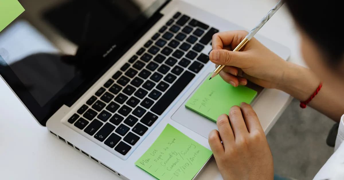 An individual writes notes on green sticky notes placed on a laptop in a work setting