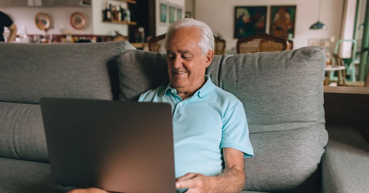 Smiling older man with white hair on a gray sofa using a laptop in a living room