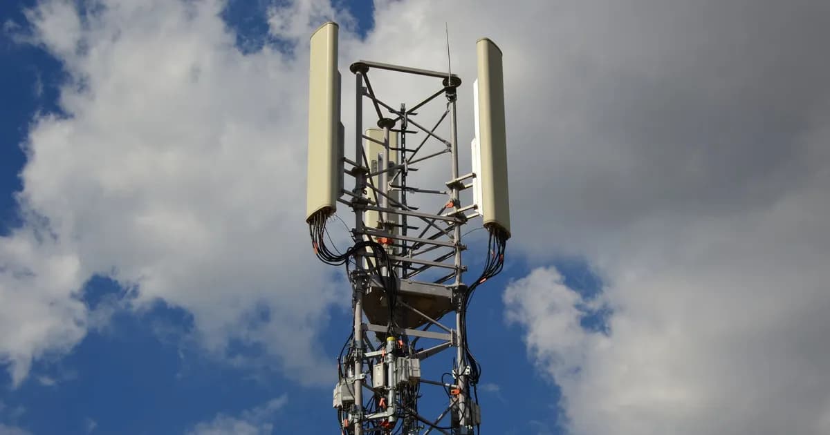 Cell tower top with large cellular antenna panels and cabling against a blue sky with clouds