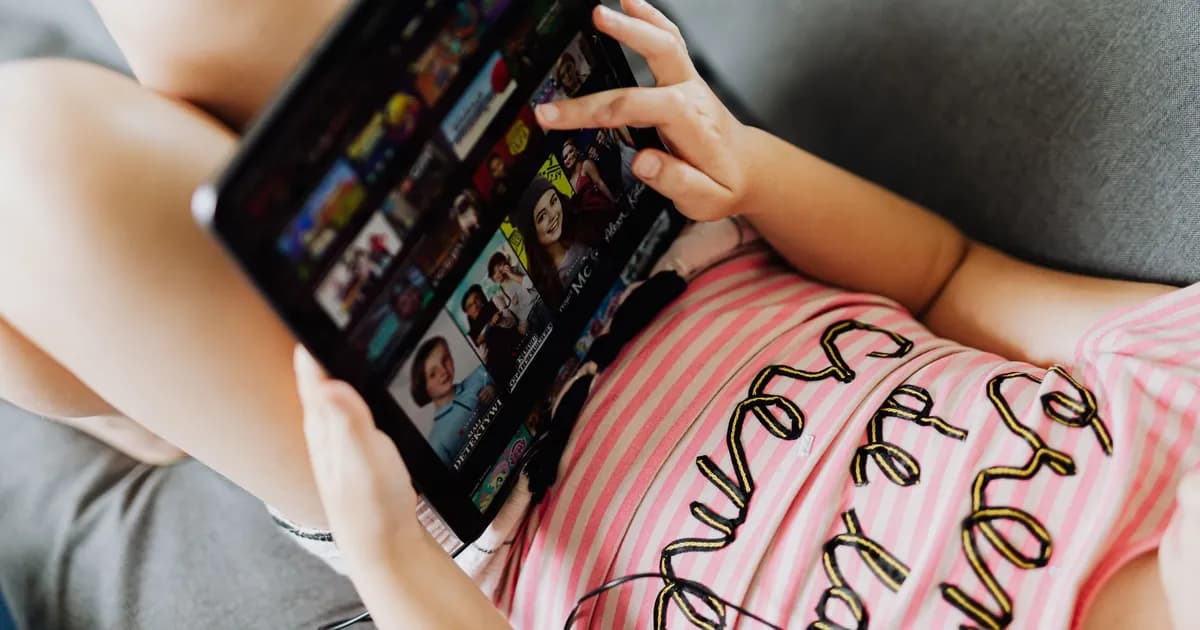 Child in a pink striped top holding a tablet showing a grid of streaming show thumbnails