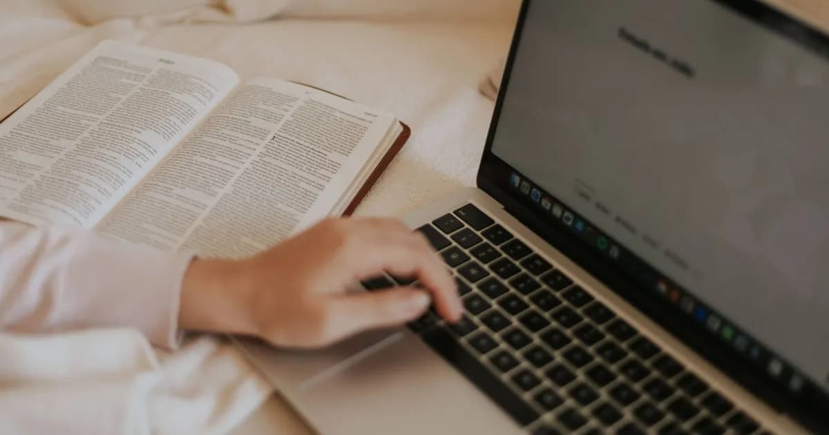 Hand on a laptop keyboard beside an open book with dense printed text