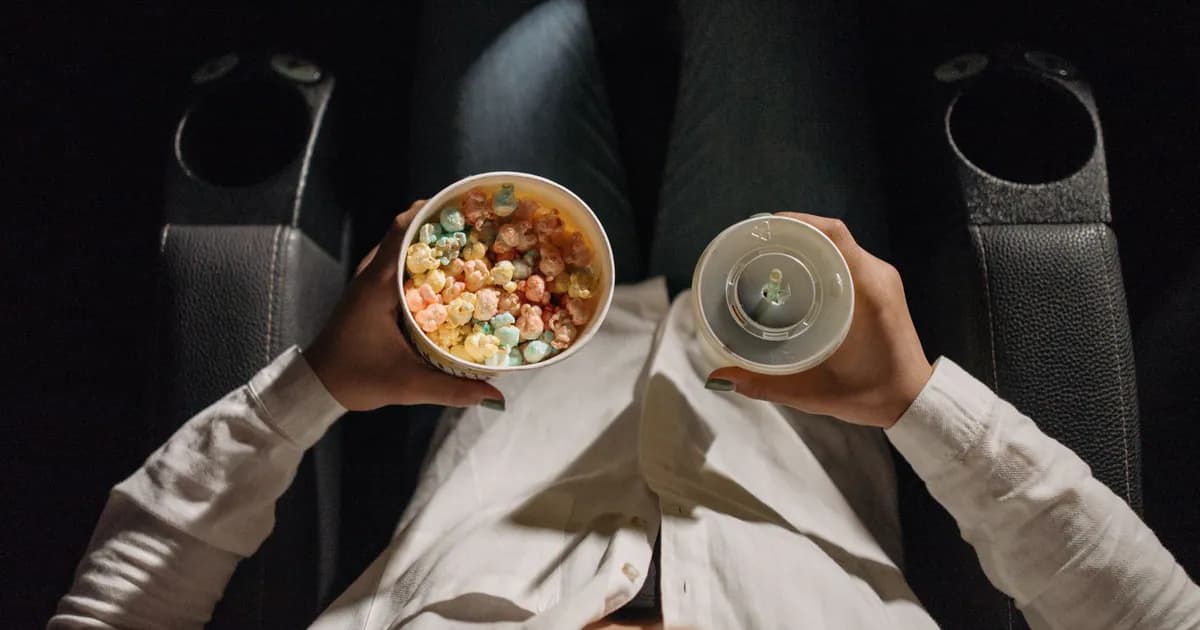 Close-up of a person holding colorful popcorn and a drink, seated in a movie theater